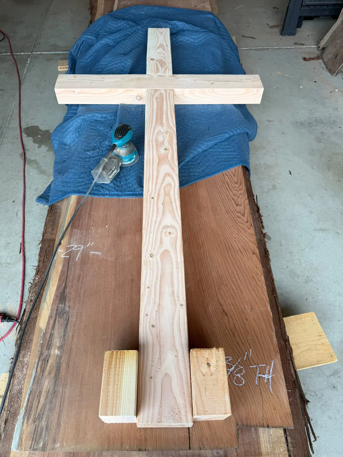 Unfinished 7-foot Douglas fir cross laid flat on a redwood slab in the shop with an orbital sander beside it