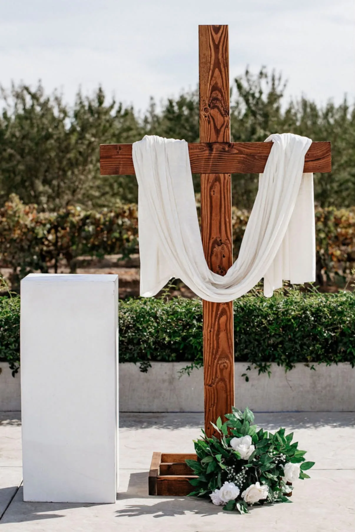 Walnut-stained 7-foot cross draped with white fabric at an outdoor wedding ceremony with white flowers at its base