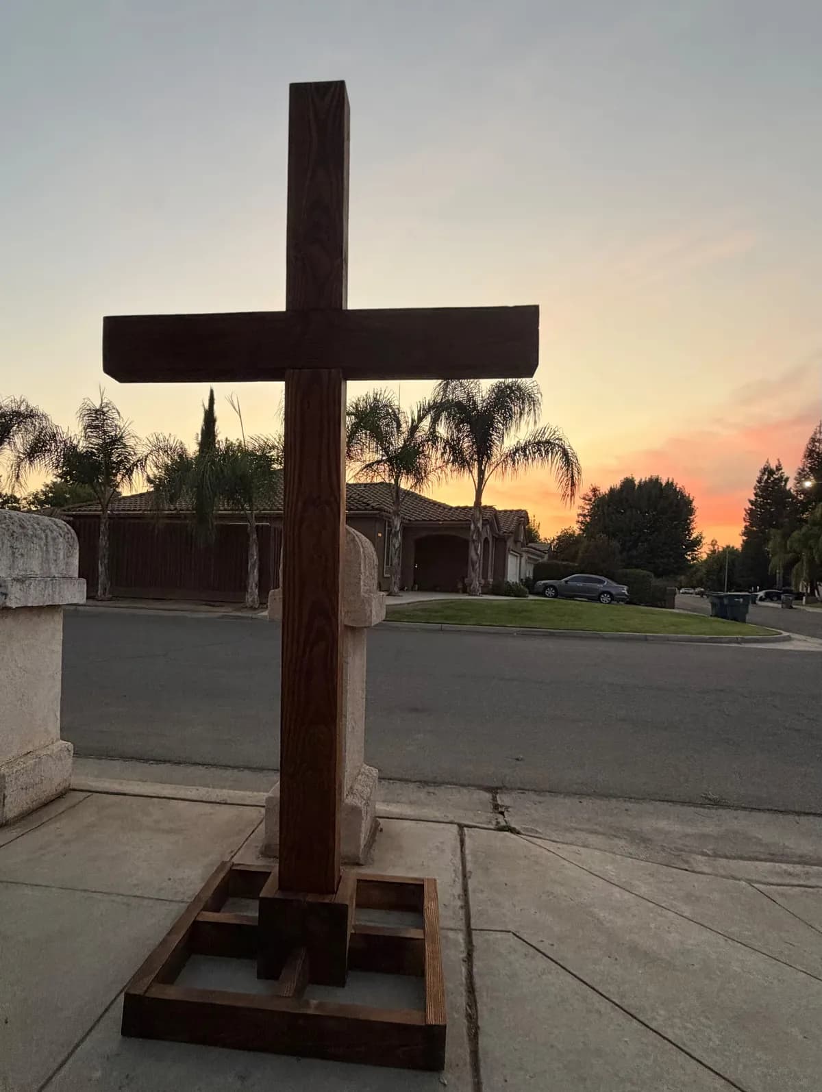 Finished walnut-stained cross standing in its wooden base on a sidewalk at sunset