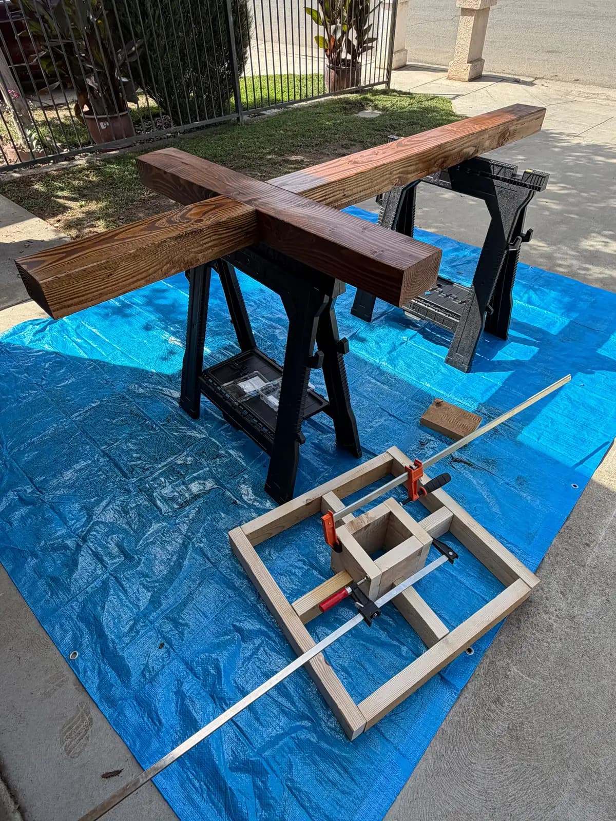 Walnut-stained Douglas fir cross resting on sawhorses beside the unfinished base clamped on a blue tarp